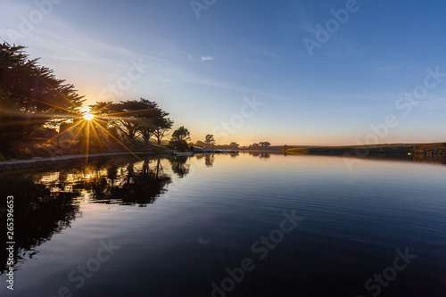 Sunrise with sun flare over river and trees silhouettes reflecting in calm water with copy space