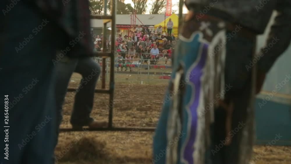A still racking focus shot of people dressed as cowboys standing aroung and talking while some people are seated in an arena waiting and watching A still racking focus shot of people dressed as cowboys standing aroung and talking while some people are seated in an arena waiting and watching