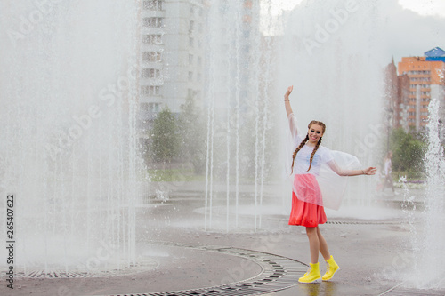 Young wet pretty girl with two braids in yellow boots stands near fountain.