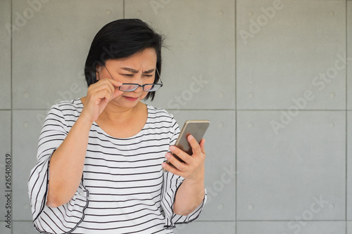 Old Asian woman holding her glasses and having difficult time looking at mobile phone device in her hand.