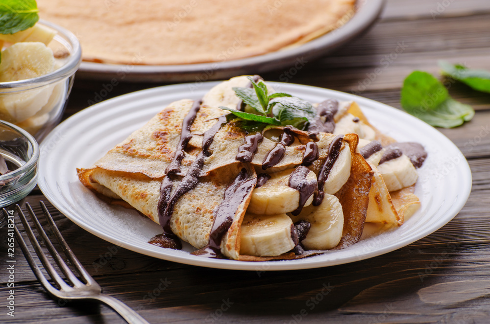 French crepes with chocolate sauce and banana in ceramic dish on wooden kitchen table French crepes with chocolate sauce and banana in ceramic dish on wooden kitchen table