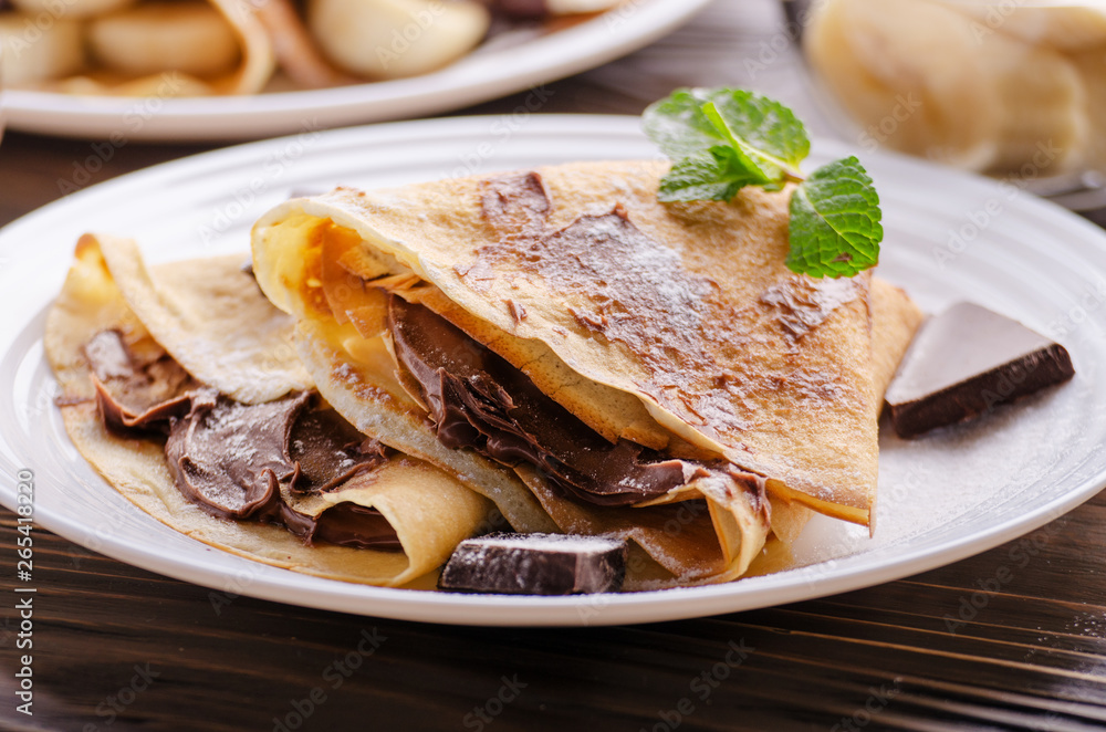 French crepes with chocolate sauce and banana in ceramic dish on wooden kitchen table
