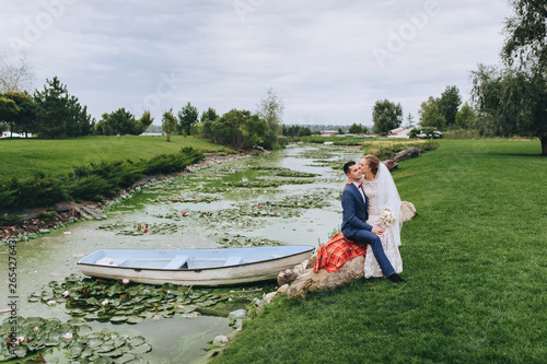 Cheerful and smiling newlyweds are hugging in the park, sitting on a stone, against the background of a boat, a river and green trees. Stylish groom and beautiful bride in nature laugh with pleasure.