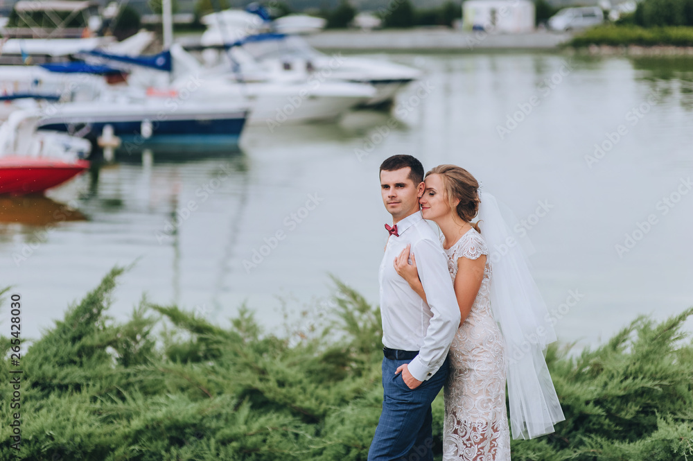 Beautiful newlyweds are hugging in the park on the green grass, on the background of yachts. Wedding portrait of a stylish groom with a bow tie and a cute curly blonde in a white dress on nature.