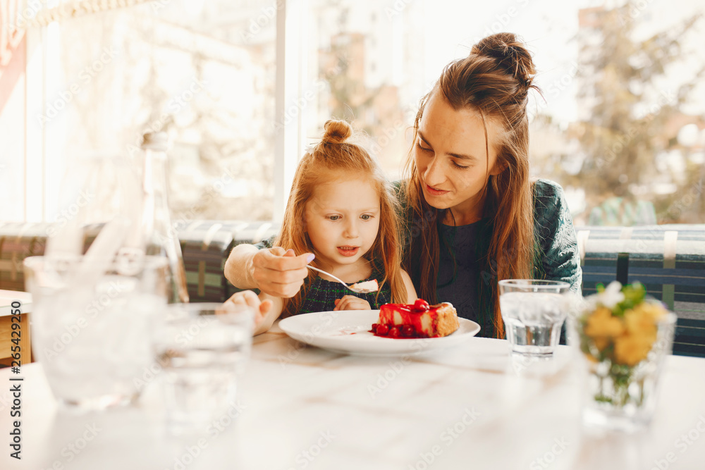 young and stylish mother with long hair and a green dress sitting with her little cute daughter in the summer cafe and she feeds her daughter with a dessert young and stylish mother with long hair and a green dress sitting with her little cute daughter in the summer cafe and she feeds her daughter with a dessert