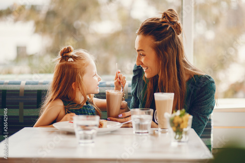 young and stylish mother with long hair and a green dress sitting with her little cute daughter in the summer cafe and she feeds her daughter with a dessert