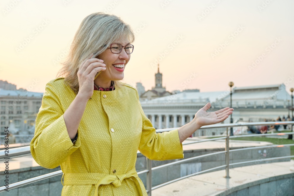 City portrait of mature smiling woman in glasses, yellow coat talking on mobile phone, background urban panorama, copy space