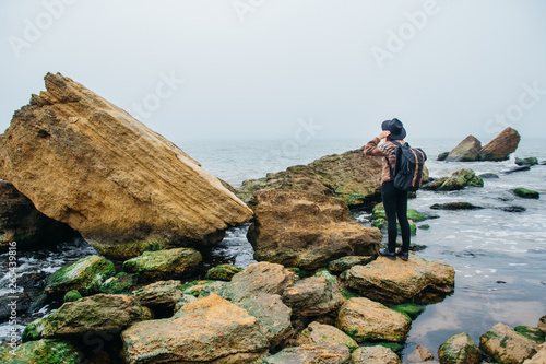 Young woman tourist in hat and with backpack on a rock against a beautiful sea, looking at sea, on coastline, on horizon. Tourism, vacation, lifestyle. Shoot from the back