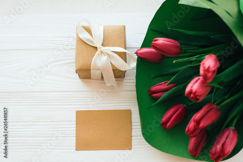 Fototapeta Naklejka Na Ścianę i Meble -  Beautiful red tulips bouquet in green paper,gift box and craft card with space for text on white wooden background flat lay. Happy mothers day. Pink tulips on white wood. Greeting card template