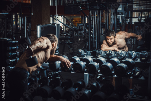 Bilde på lerret Good looking young man bodybuilder doing triceps kickback lifting dumbbells in front of the mirror looking at himself