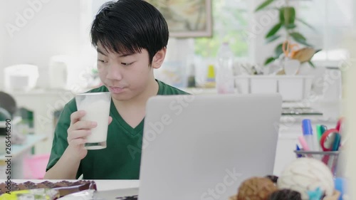 Asian young boy using scrolls through smartphone applications, while slowly eats organic milk breakfast with bread biscuit and chocolate at home.