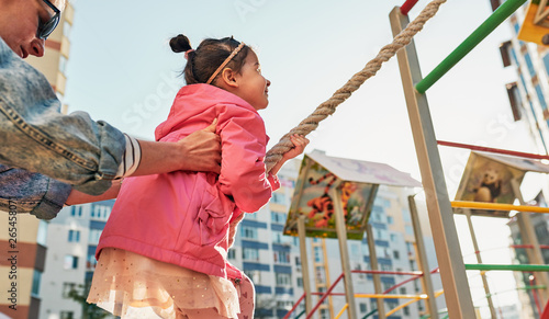 Side view image of active cute little girl climbing on a rope at playground. Mother helping her daughter to climb on a wall with a rope outside. Motherhood and childhood concept. Lifestyle, education.