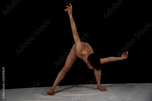 Male body-colored ballet dancer practicing in the studio with hands and legs on a black background. The attractive young man shows modern dance poses.