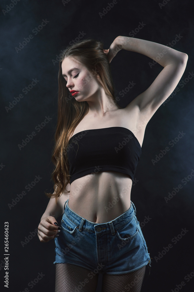 Young beautiful, slender, passionate brown-haired woman with bright red lips and long hair in a black top and denim shorts on a dark background Young beautiful, slender, passionate brown-haired woman with bright red lips and long hair in a black top and denim shorts on a dark background