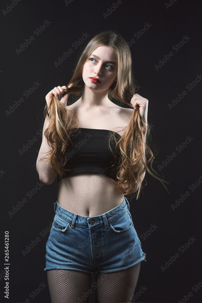 Young beautiful, slender, passionate brown-haired woman with bright red lips and long hair in a black top and denim shorts on a dark background Young beautiful, slender, passionate brown-haired woman with bright red lips and long hair in a black top and denim shorts on a dark background