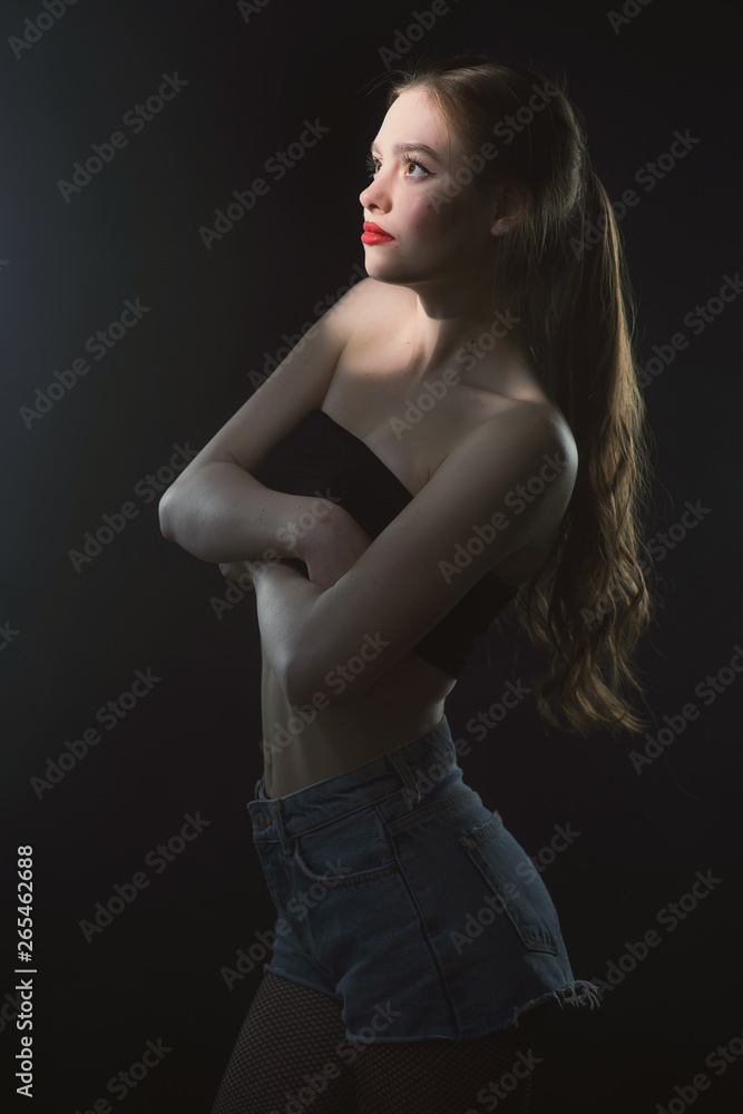 Young beautiful, slender, passionate brown-haired woman with bright red lips and long hair in a black top and denim shorts on a dark background Young beautiful, slender, passionate brown-haired woman with bright red lips and long hair in a black top and denim shorts on a dark background