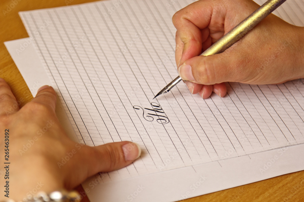 female hand writes with an ink pen on a white paper sheet with stripes. stationery on wooden desk close up top view. spelling lessons and caligraphy exercises female hand writes with an ink pen on a white paper sheet with stripes. stationery on wooden desk close up top view. spelling lessons and caligraphy exercises