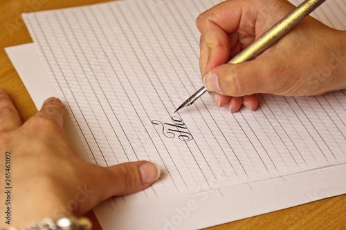 female hand writes with an ink pen on a white paper sheet with stripes. stationery on wooden desk close up top view. spelling lessons and caligraphy exercises