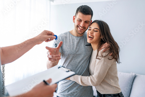 Happy couple is taking keys from their new house from broker and smiling. Hands of estate agent giving keys to the couple. The agent handed the keys a young couple photo