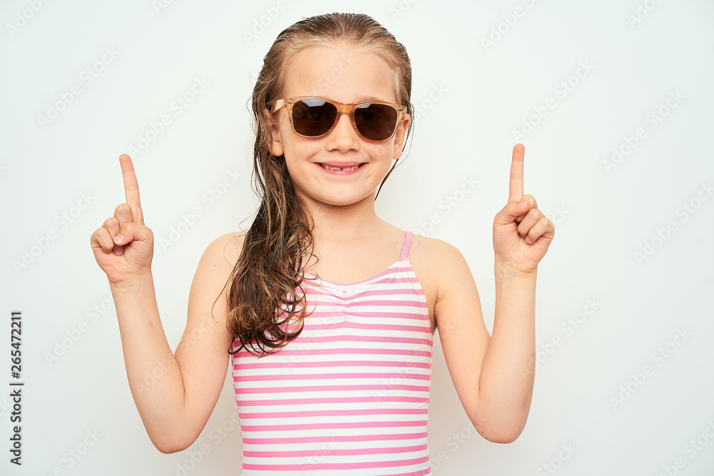 Smiling little preschool girl with wet hair photographed against white background wearing swimsuit and sunglasses pointing up with finger towards empty space