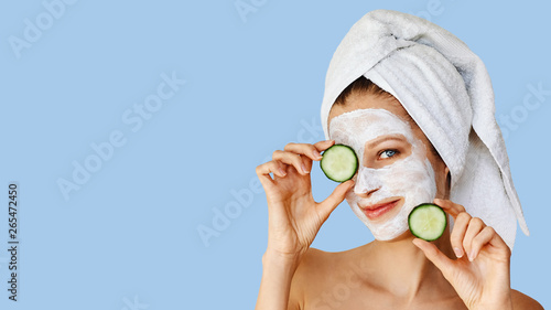 Beautiful young woman with facial mask on her face holding slices of cucumber. Skin care and treatment, spa, natural beauty and cosmetology concept.