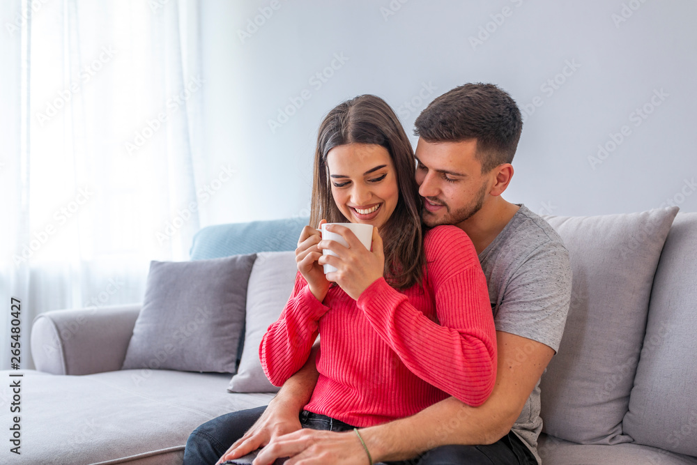 Beautiful young couple is talking and smiling while sitting on sofa at home. Woman is holding a cup. Cheerful beautiful young couple sitting and hugging on sofa at home Beautiful young couple is talking and smiling while sitting on sofa at home. Woman is holding a cup. Cheerful beautiful young couple sitting and hugging on sofa at home
