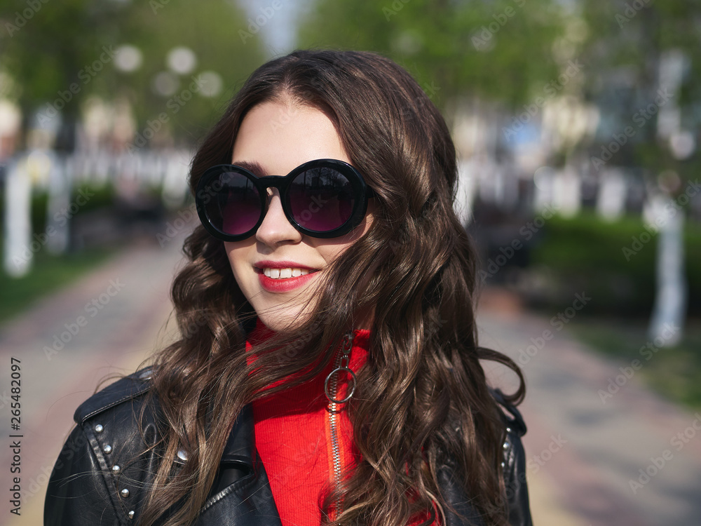 Headshot of young happy fashionable elegant brown-haired woman in sunglasses with long curly hair enjoying spring warm weather at city street blurring background. Headshot of young happy fashionable elegant brown-haired woman in sunglasses with long curly hair enjoying spring warm weather at city street blurring background.