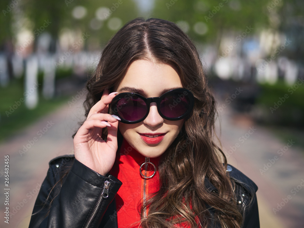 Headshot of young happy fashionable elegant brown-haired woman in sunglasses with long curly hair enjoying spring warm weather at city street blurring background. Headshot of young happy fashionable elegant brown-haired woman in sunglasses with long curly hair enjoying spring warm weather at city street blurring background.