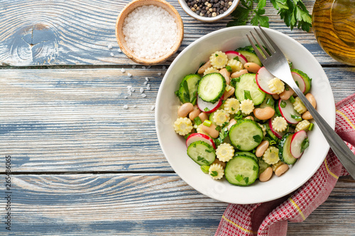 Healthy vegetable salad with cucumber, radish, beans, mini corn and herbs on rustic wooden background. Top view. Copy space.