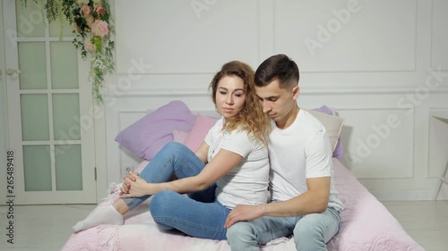 Young couple in love man and woman in white t-shirts and jeans are sitting on bed in the bedroom, man is looking at woman, but she is looking at camera all time.