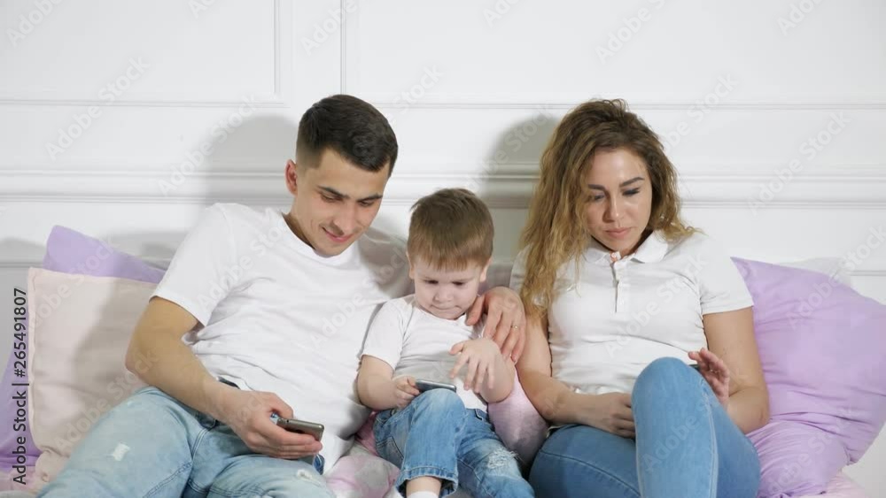 Family portrait: mom, dad and son are looking in their mobile phones laying on the bed, family look style. Gadgets addiction. Family portrait: mom, dad and son are looking in their mobile phones laying on the bed, family look style. Gadgets addiction.