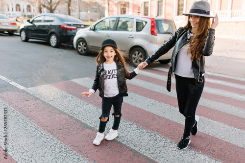 Stylish little girl in black hat stands at an crossroad next to her mother and look with admiration with cars on background. Slim trendy young woman passes long-haired daughter across the road.