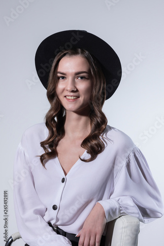 Portrait of a young girl with long hair in a white blouse and a black hat made in the studio on a light background.
