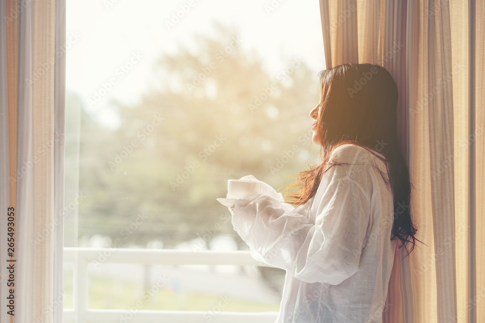 Asian woman in bedroom drinking coffee after wake up near window, sunny morning.  Lifestyle Concept.