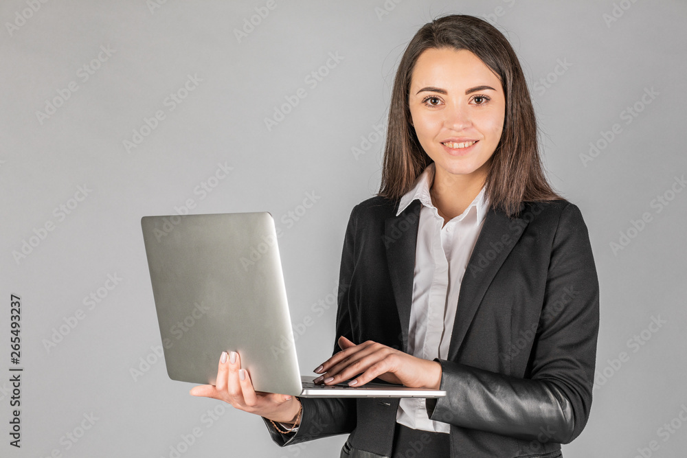 Photo of a young cheerful business woman standing over a gray wall with a laptop Photo of a young cheerful business woman standing over a gray wall with a laptop