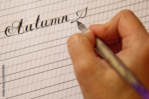 female hand writes with the inky pen the word autumn on a white paper sheet with stripes. stationery on desk close up top view. spelling lessons and caligraphy exercises. Template, layout, background