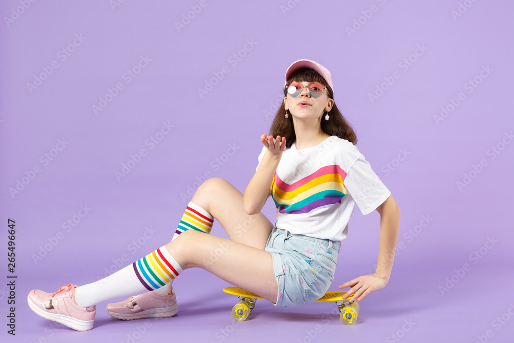 Naklejka premium Tender teen girl in vivid clothes, eyeglasses sitting on yellow skateboard blowing sending air kiss isolated on violet pastel background. People sincere emotions lifestyle concept. Mock up copy space.