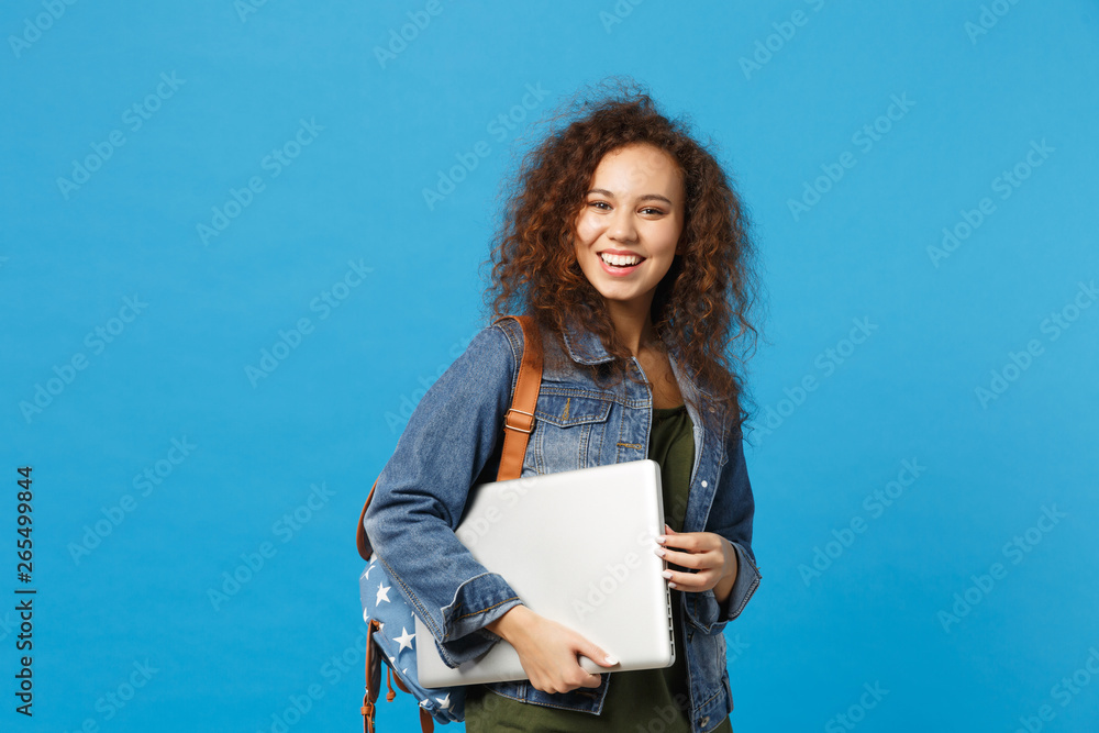 Young african american girl teen student in denim clothes, backpack hold pc isolated on blue wall background studio portrait. Education in high school university college concept. Mock up copy space.