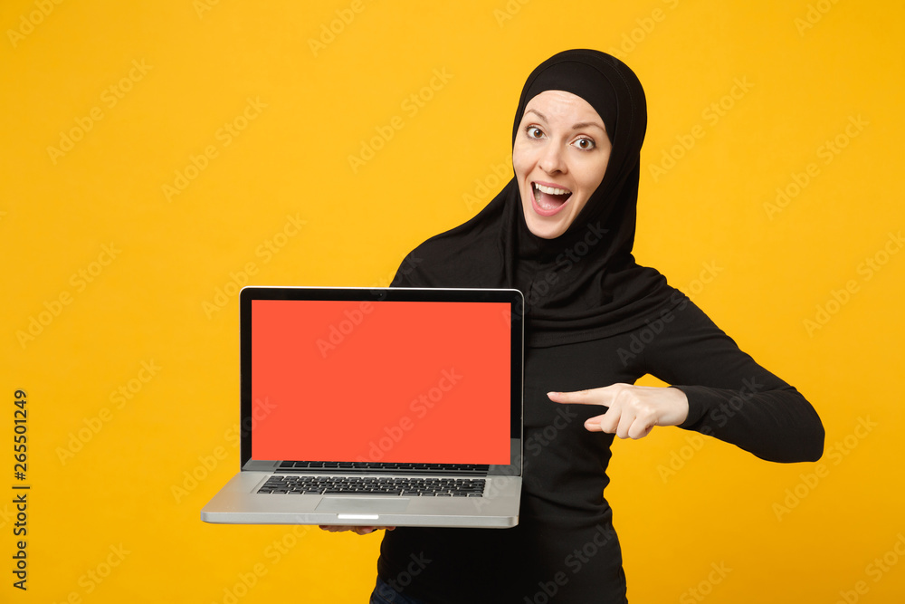Young arabian muslim employee woman in hijab black clothes hold laptop pc with blank empty screen isolated on yellow wall background, studio portrait. People lifestyle concept. Mock up copy space. Young arabian muslim employee woman in hijab black clothes hold laptop pc with blank empty screen isolated on yellow wall background, studio portrait. People lifestyle concept. Mock up copy space.