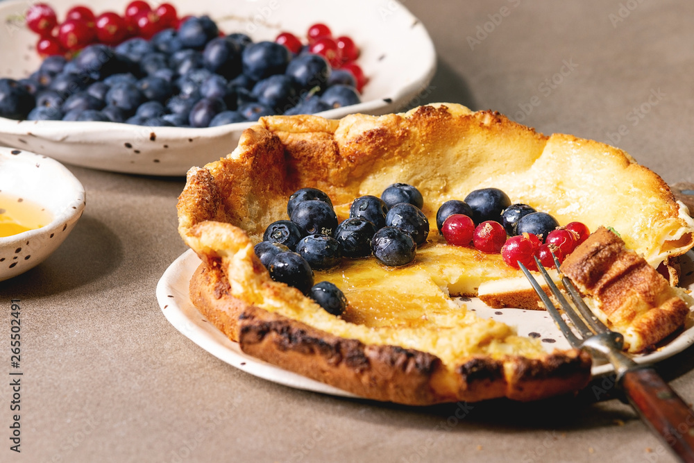 Started eaten fresh baked Dutch baby pancake in ceramic plate with blackberry and red currant berries, bowl of honey, jug of cream, vintage cutlery over beige kitchen table. Close up Started eaten fresh baked Dutch baby pancake in ceramic plate with blackberry and red currant berries, bowl of honey, jug of cream, vintage cutlery over beige kitchen table. Close up