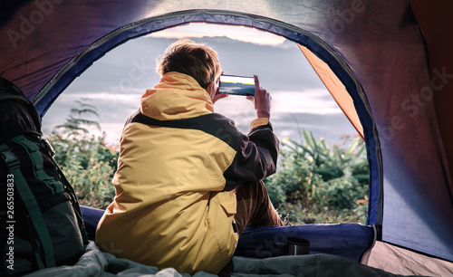 Young male hiker using mobile phone taking a photo with inside a tent in morning enjoying the leisure and freedom.
