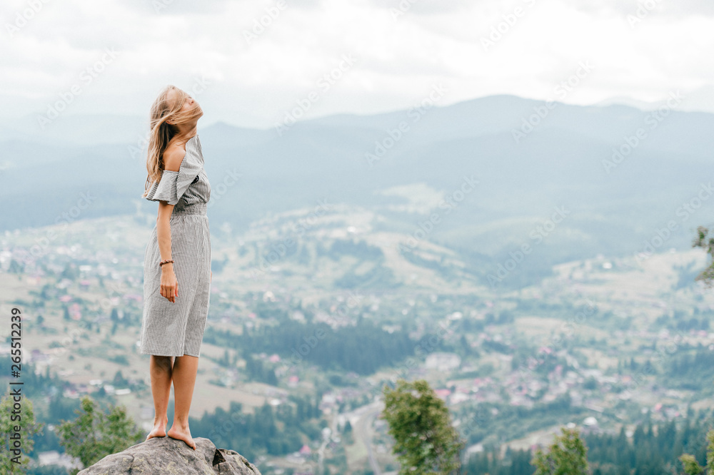 Naklejka premium Young beautiful barefoot blonde girl with long hair in summer dress standing on top of conquered mountain at stone and enjoying fabulous landscape scenic view with mountains and village in valley