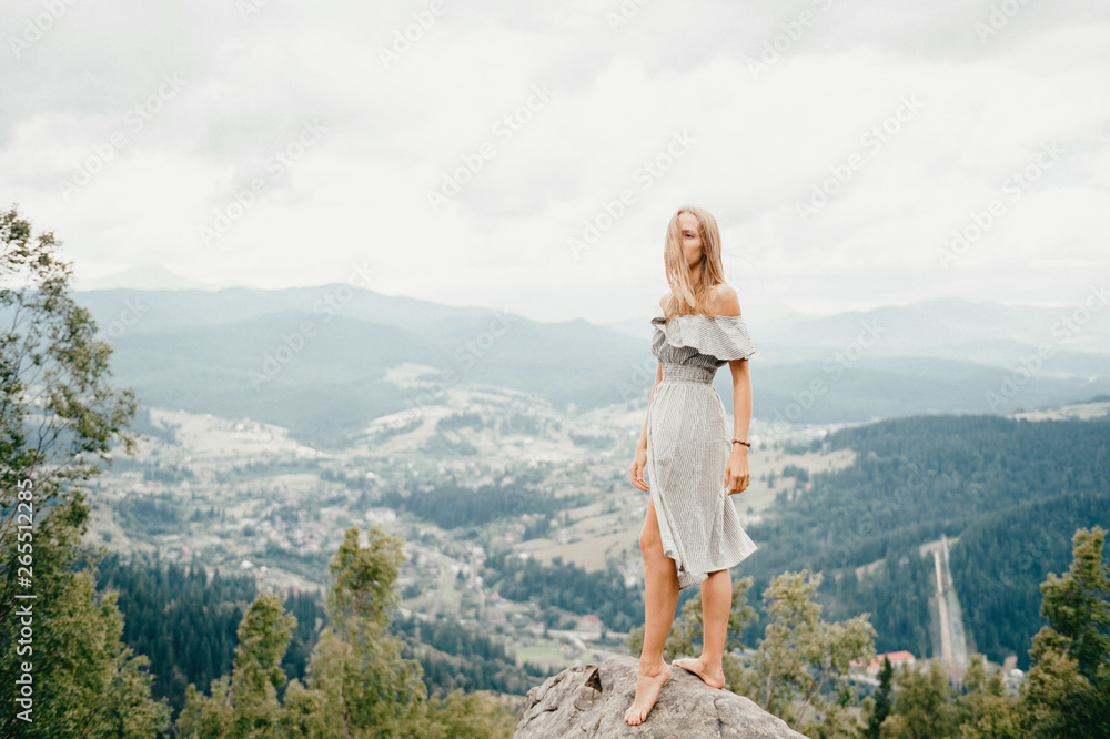 Naklejka premium Young beautiful barefoot blonde girl with long hair in summer dress standing on top of conquered mountain at stone and enjoying fabulous landscape scenic view with mountains and village in valley