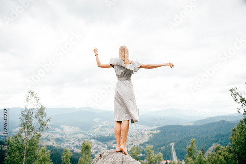 Young beautiful barefoot blonde girl with long hair in summer dress standing on top of conquered mountain at stone and enjoying fabulous landscape scenic view with mountains and village in valley