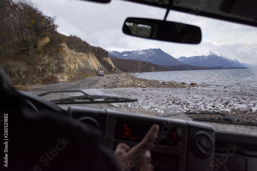 Inside view from the interior of a vehicle to the front window, with a dirt rocky route on the background next to a lake. The driver is in a blur pointing out and the interior is dark, with copy space