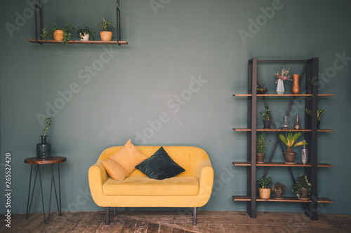 Interior design room or studio. Furniture against a green wall and wooden floor - a wooden table with a vase, a yellow sofa with cushions, a wardrobe with flowers and plants, a hanging shelf