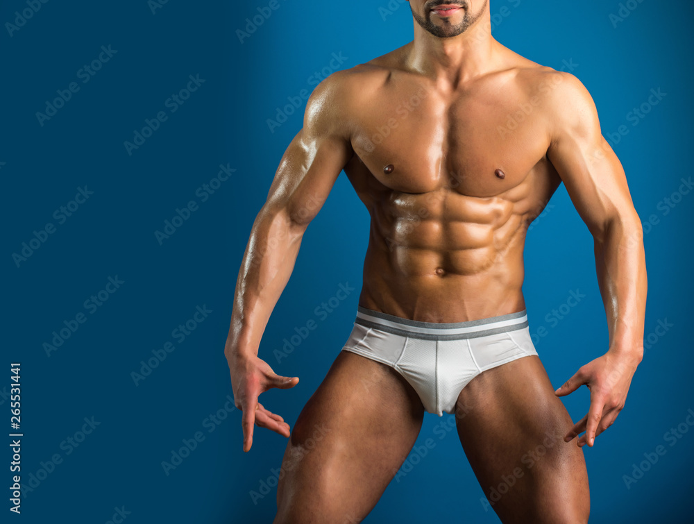 Perfect fit body of young handsome guy isolated on blue background. Young male model pose for gym or competition advertising campaign. Close up of strong muscular mans body in underwear.