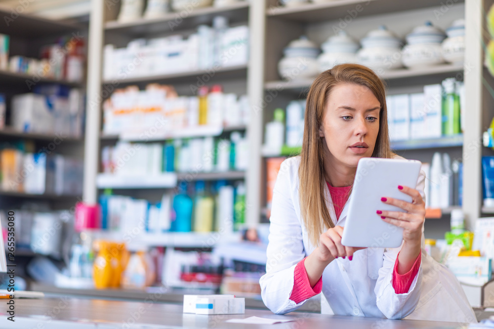 Young pharmacist holding a tablet and box of medications. Young positive brunette pharmacist woman in drug store with tablet pc. Female pharmacist with digital tablet searching for medication