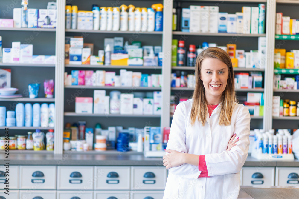 Portrait of a smiling pharmacist with arms crossed at modern pharmacy. Beautiful woman wearing in white lab coat working in drugstore.
