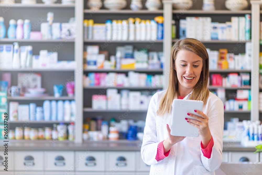 Smiling pharmacist using a tablet-pc scrolling with her finger on the touchscreen while standing in front of shelves of medication. Pharmacist using digital tablet in pharmacy